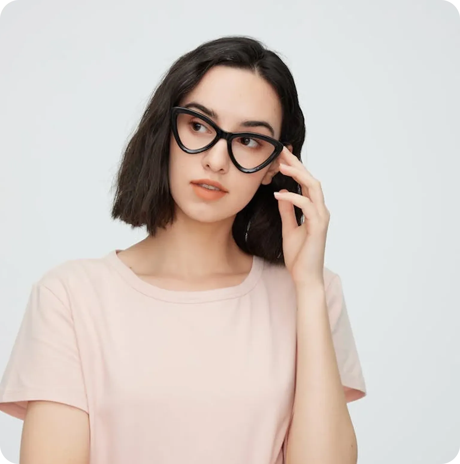 Young woman with short dark hair wearing black cat-eye glasses and a light pink t-shirt touching her glasses.